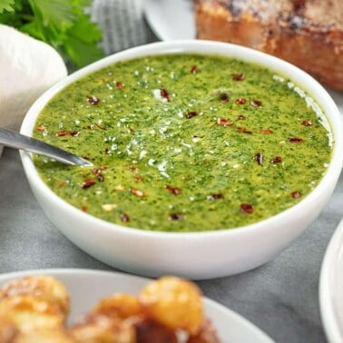 Angled view of Chimichurri sauce in a white bowl surrounded by some other foods on the table.