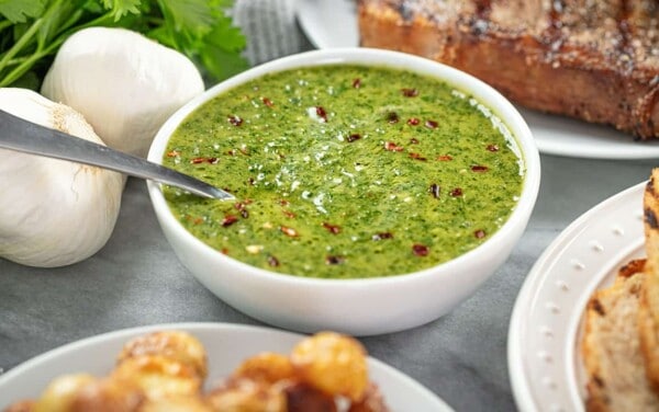 Angled view of Chimichurri sauce in a white bowl surrounded by some other foods on the table.