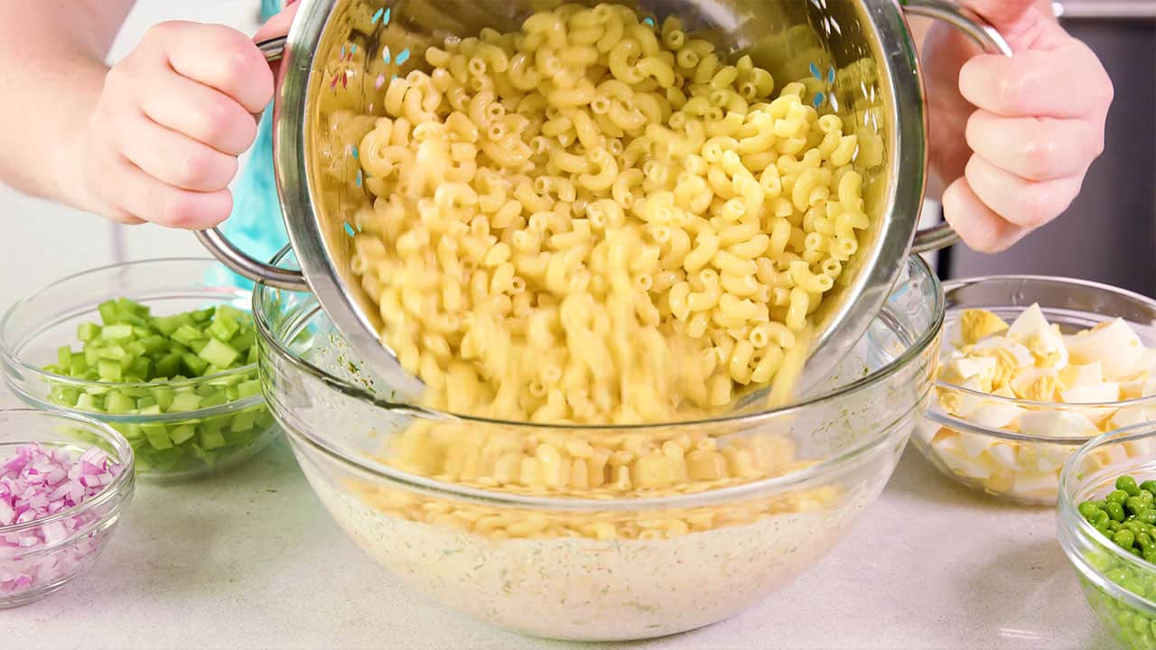 Angled view of a large stainless steel colander pouring elbow macaroni pasta into a large clear glass mixing bowl filled with salad dressing ingredients.