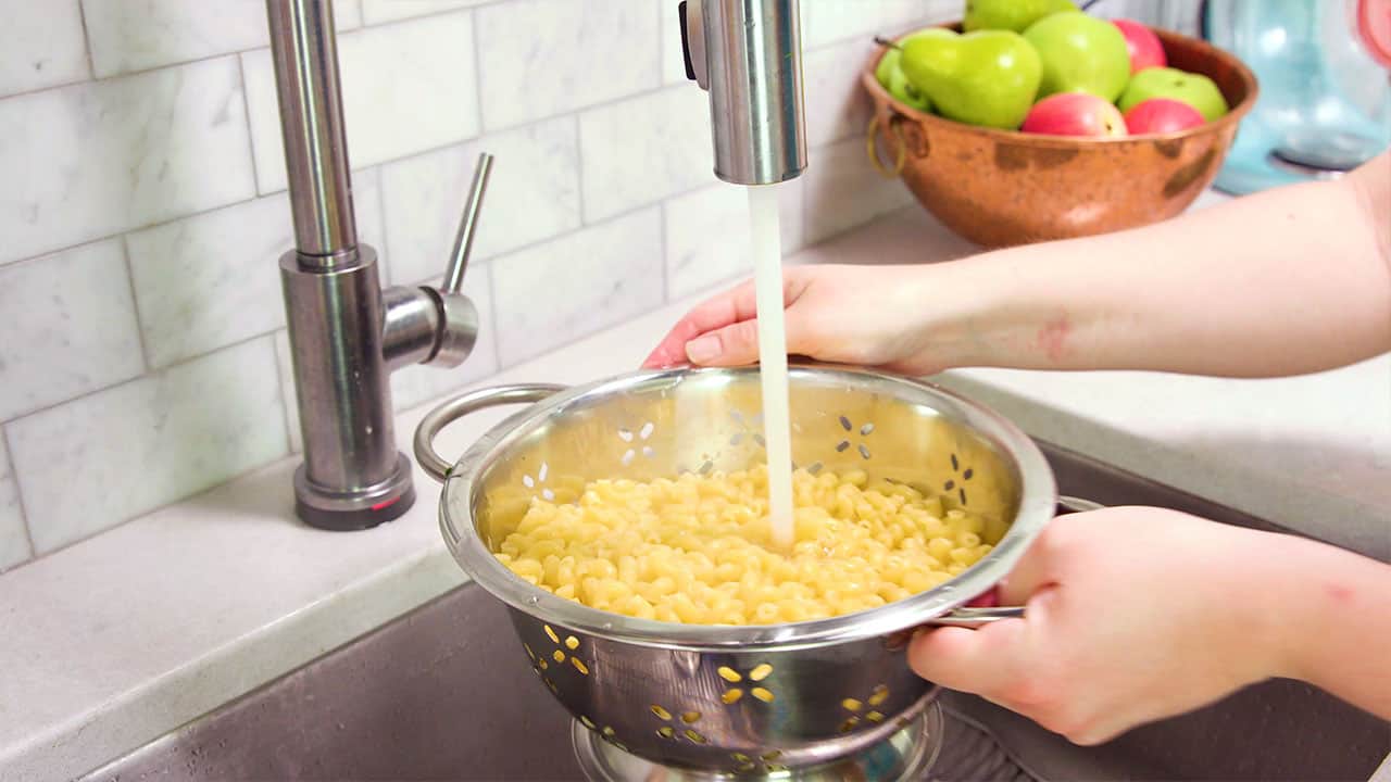 Angled view of a large stainless steel colander filled with elbow macaroni under the running water of a faucet to rinse and cool the pasta.