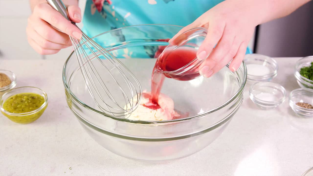 Angled view of large clear glass mixing bowl with a small clear glass mixing bowl pouring red wine vinegar with other ingredients for the salad dressing and a wire whisk at the ready for stirring.