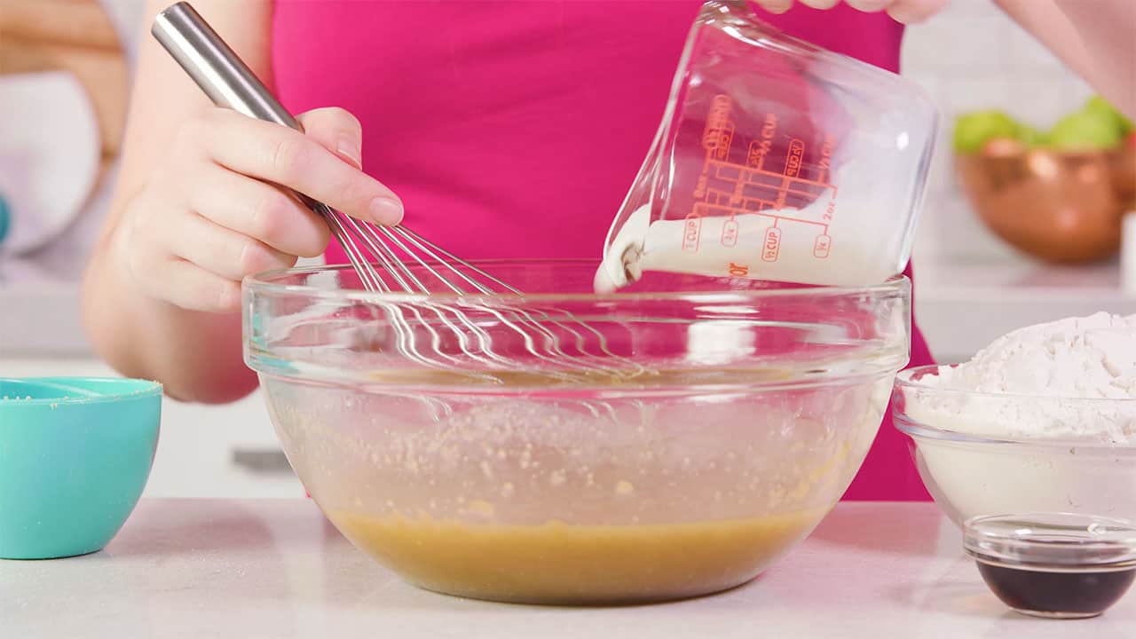 Side view of large clear glass mixing bowl with a clear glass measuring cup pouting heavy cream and a wire whisk at the ready for stirring.