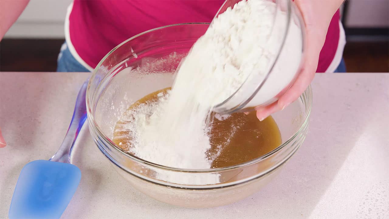 Angled view of large glass mixing bowl with wet ingredients combined with dry ingredients from a smaller clear glass mixing bowl and a plastic spatula at the ready for stirring.