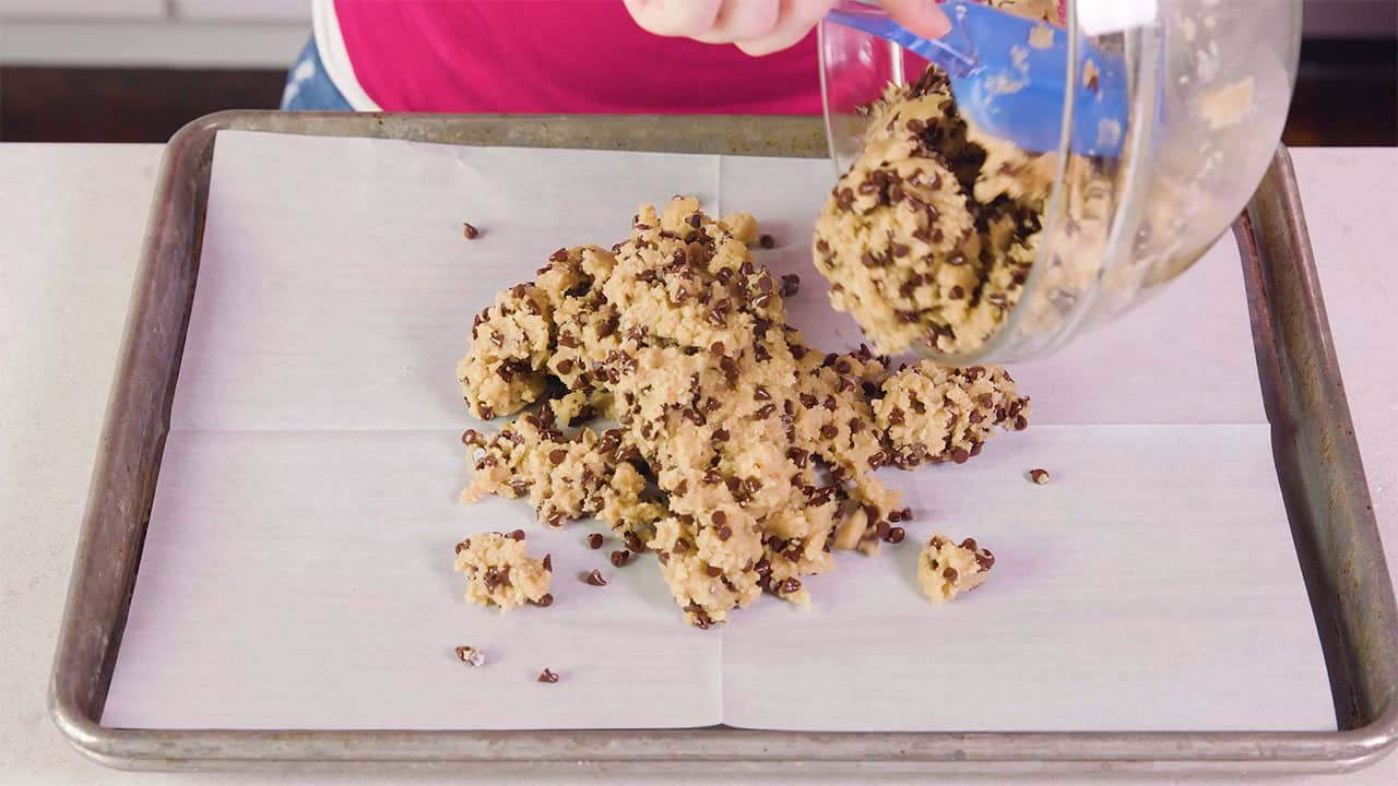 Angled view of metal baking sheet lined with parchment paper and chocolate chip cookie dough being poured from a clear glass mixing bowl and scooped out with a plastic spatula.