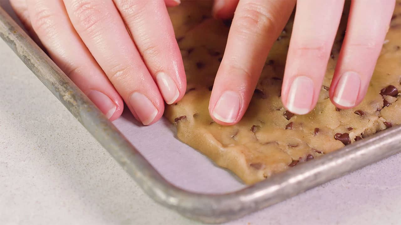 Close-up view of the corner of a metal baking sheet lined with parchment paper showing a detailed view of fingertips forming the chocolate chip cookie dough into a flat sheet and straight edges.