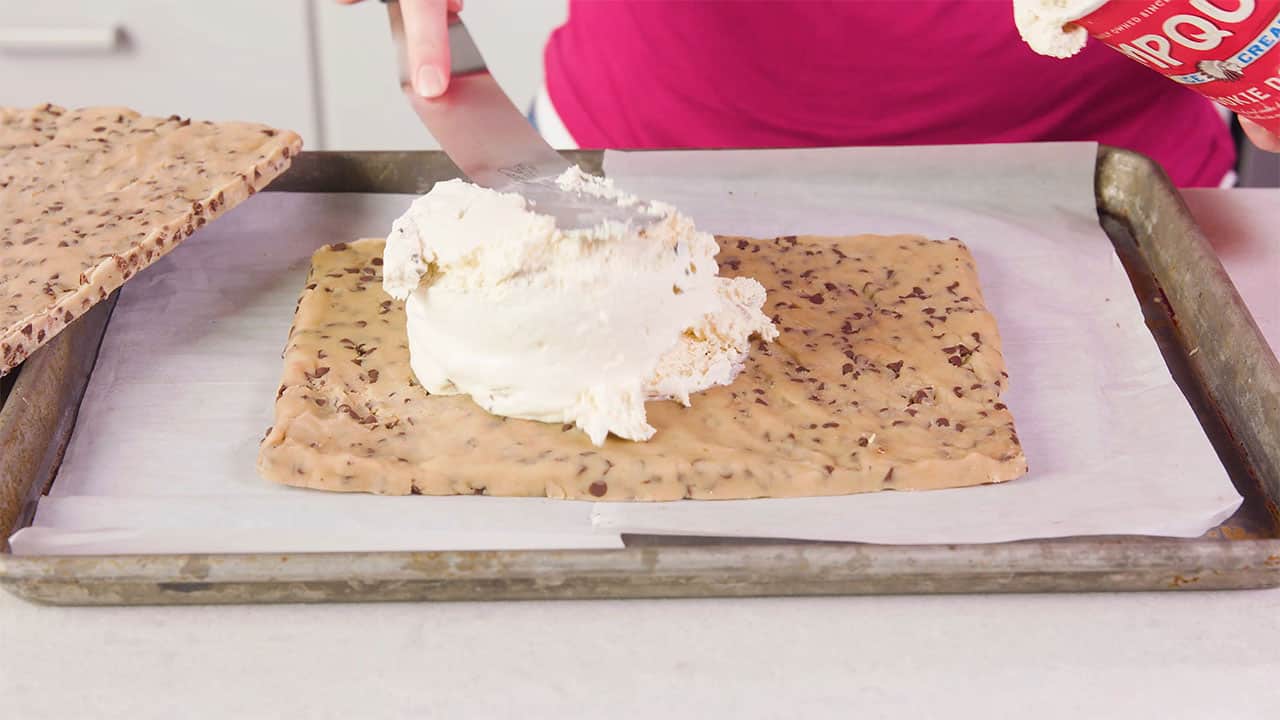 Angled view of metal baking sheet lined with parchment paper with a formed sheet of chocolate chip cookie dough and vanilla ice cream placed on top and formed as the inside layer with a flat metal spatula and a second sheet of cookie dough to the side ready to go on as the top layer of the sandwich.