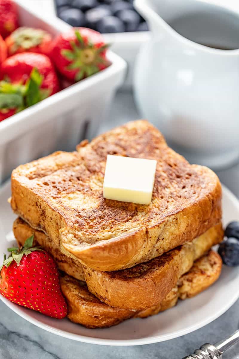 A stack of French Toast with a pad of butter on it all on a white plate with some berries on it.
