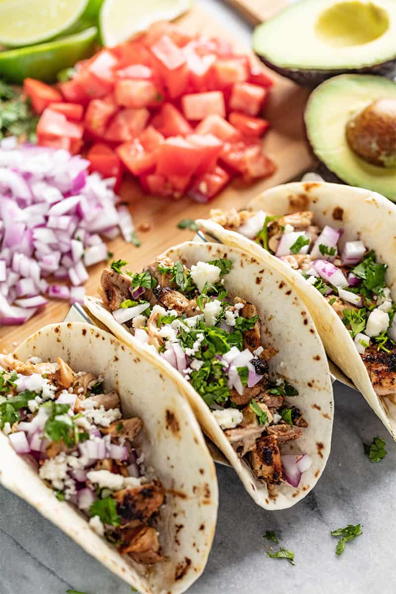 Tacos de frango num suporte de tacos com os seus ingredientes ao fundo.