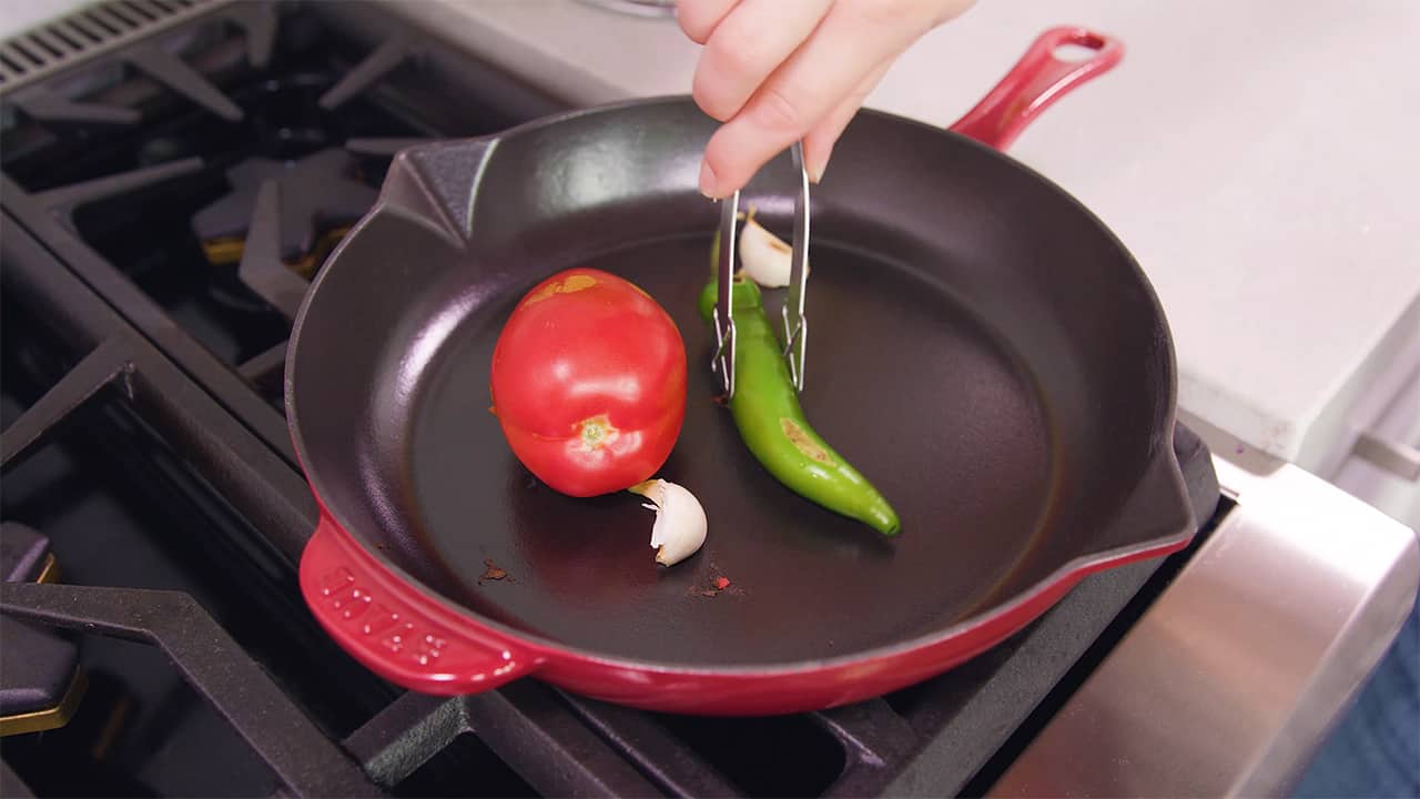 Using a skillet on a stove-top, char the outside of one Roma tomato, one green Serrano pepper, and two cloves of garlic, using small metal tongs to move and remove from skillet.