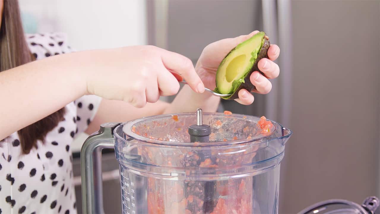 Using a spoon, remove a halved avocado from its skin and add to a food processor.