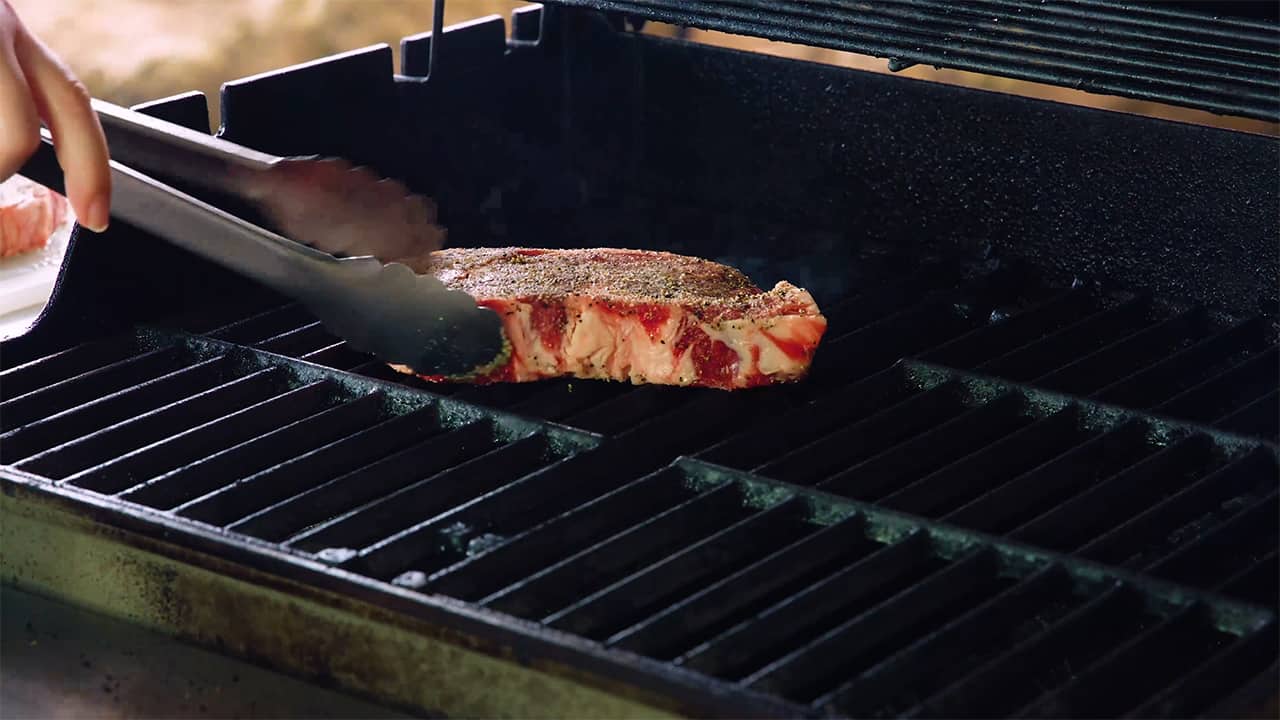 Using metal tongs, place the raw steak on the hot grill.