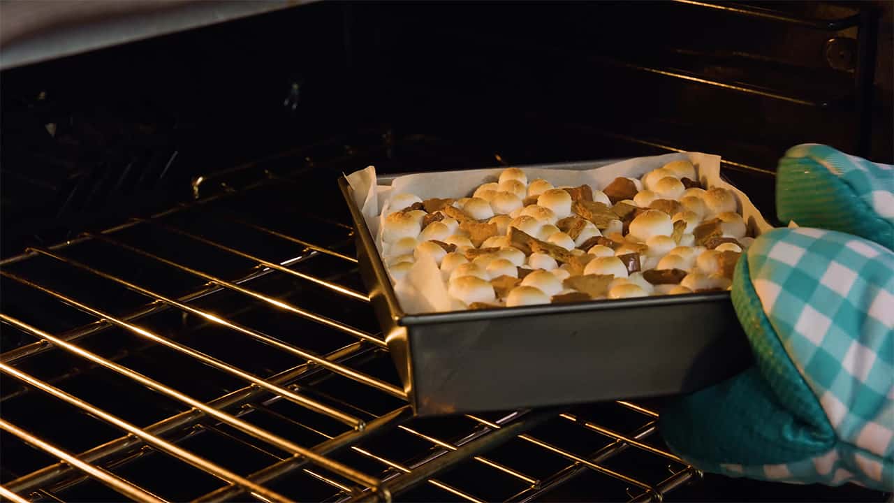 Angled view of square metal brownie pan lined with parchment paper being pulled from an open oven and showing the golden brown toasted marshmallows on the top layer of the brownies.