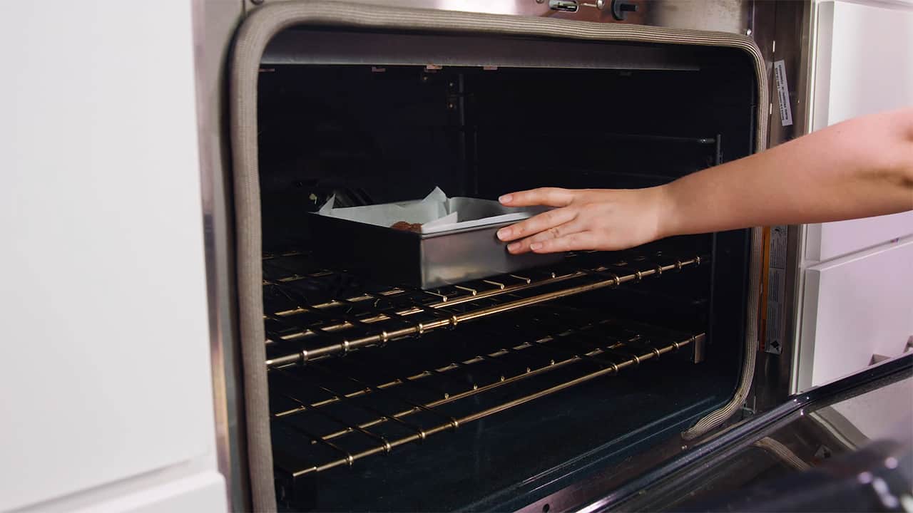 Angled view of an open oven with a square metal brownie pan lined with parchment paper and filled with brownie mix placed on a metal rack in the middle of the oven.