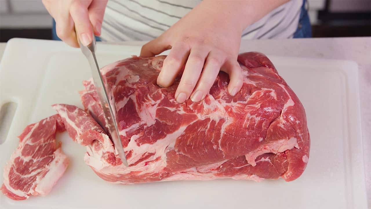 Overhead view of raw beef being sliced by a kitchen knife on a white cutting board.
