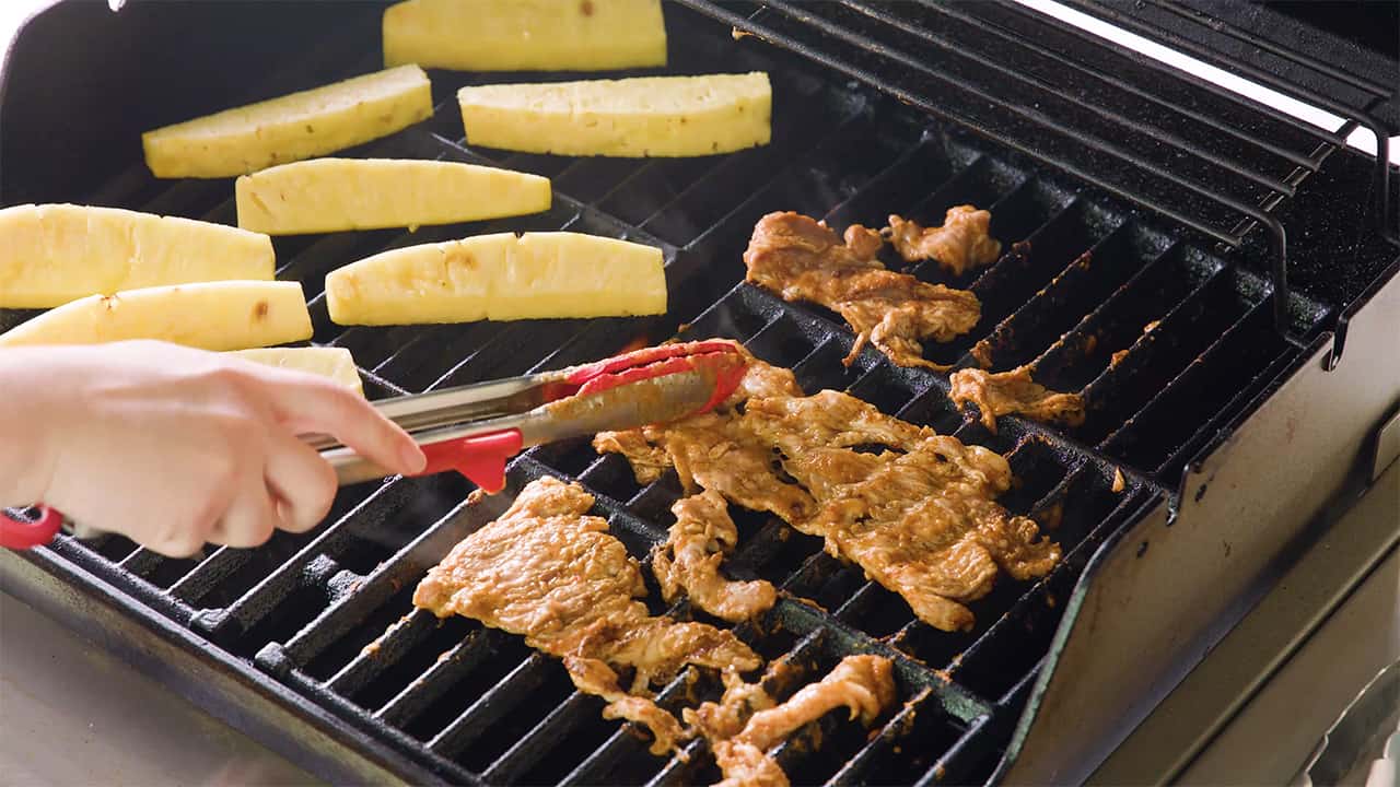 Angled view of open grill with grilling marinaded beef in the foreground and fresh pineapple spears on the left side of the grill.