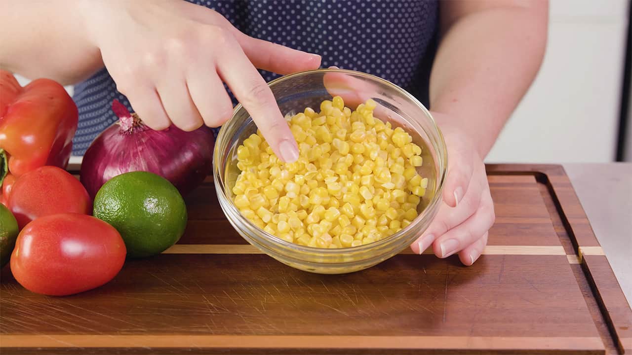 Angled view of medium sized clear glass mixing bowl filled with yellow sweet corn and other ingredients to the side all on a wood cutting board.