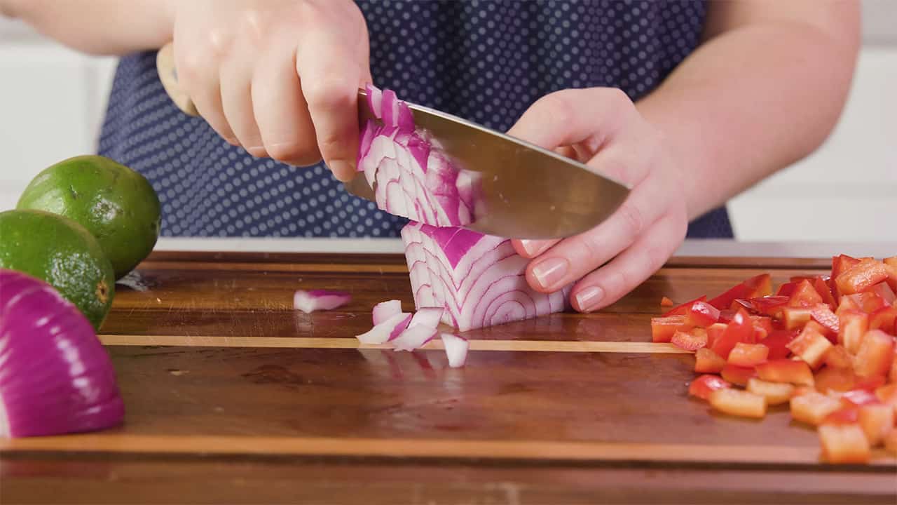 Side view of a red onion being diced with diced tomato to the right and fresh uncut limes to the left all on a wood cutting board.