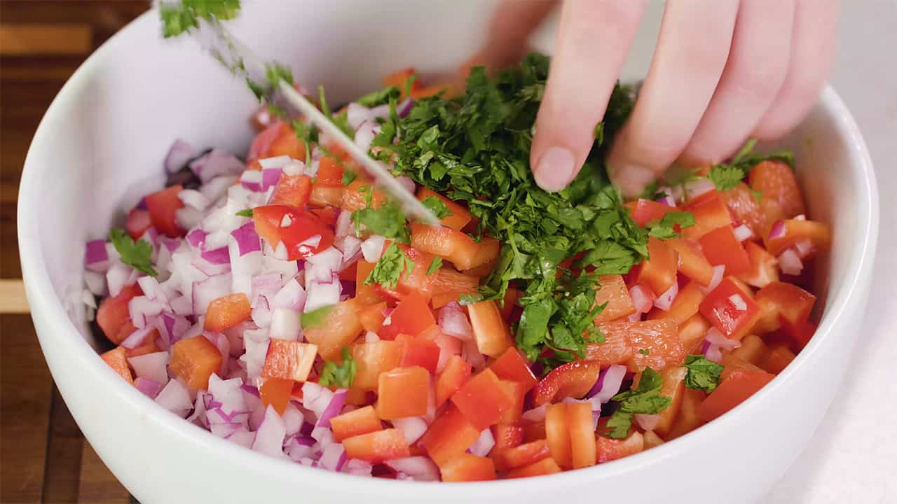 Close-up view of a large white mixing bowl filled with diced vegetables for the salad being topped with chopped cilantro.