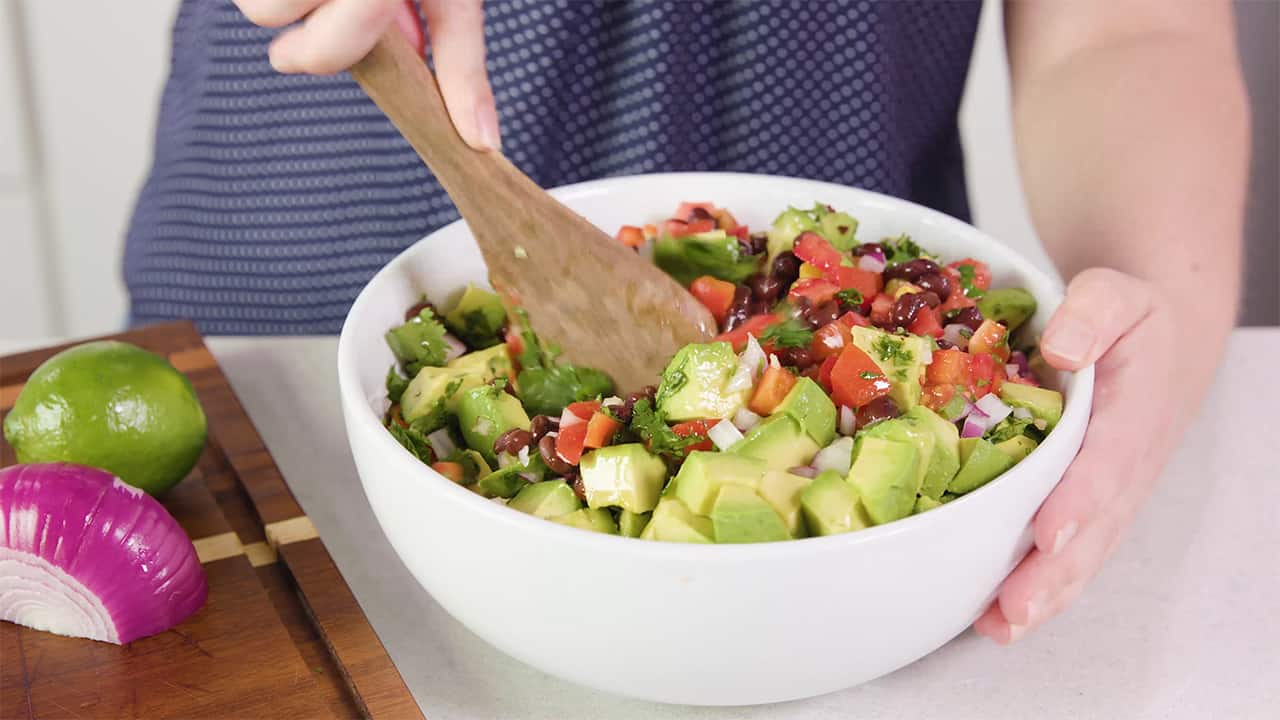Angled view of a large white glass mixing bowl filled with all the fresh ingredients for black bean salad and being mixed with a wooden spoon to combine.