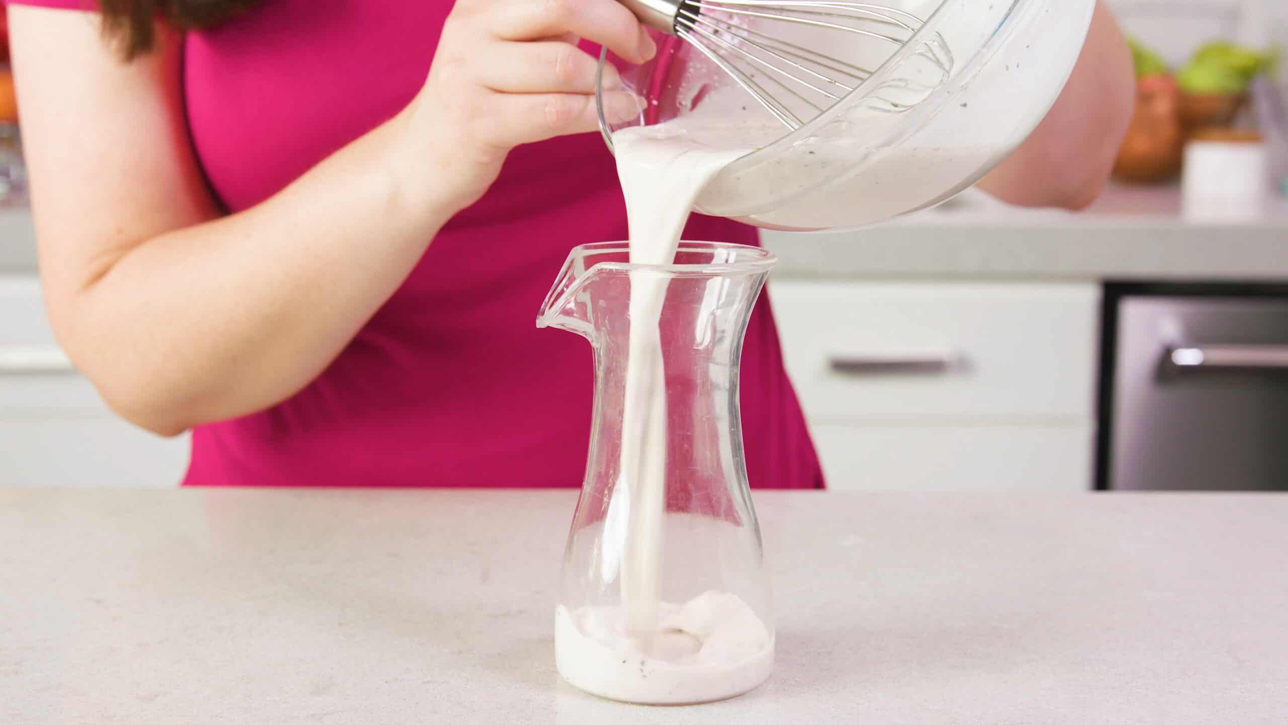 Side view of clear plastic salad dressing vessel with salad dressing being poured from a large clear glass mixing bowl.