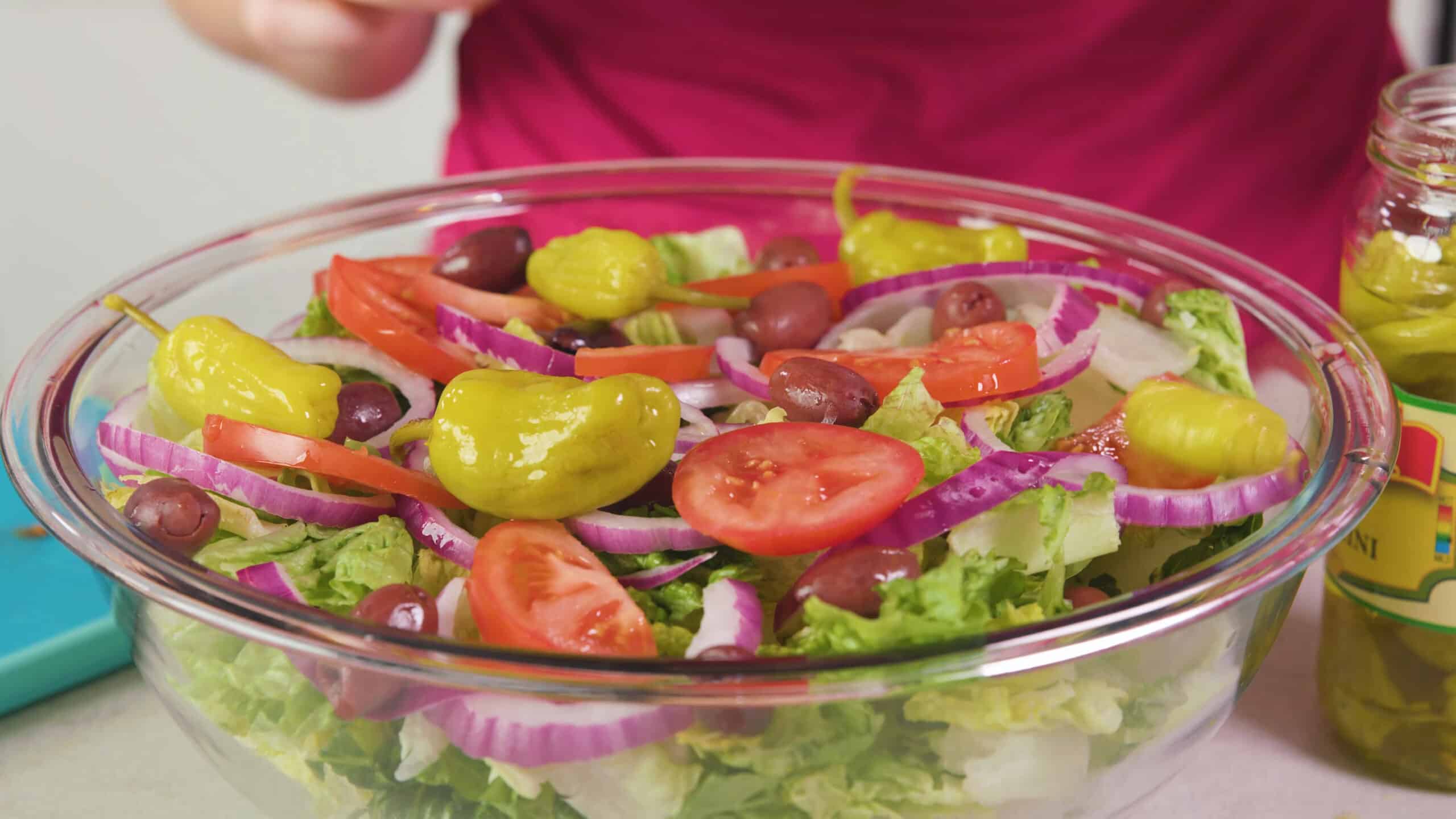 Angled view of a large clear glass mixing bowl filled with salad ingredients topped with pepperoncinis, pitted kalamata olives, sliced red onion, and sliced tomatoes.