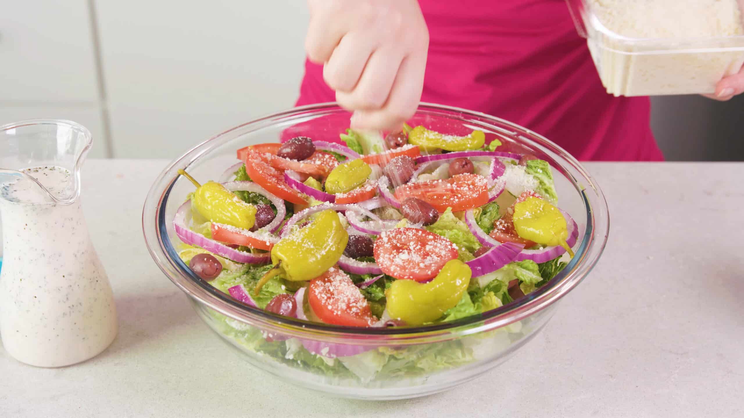Angled view of a large clear glass mixing bowl filled with fresh green salad and being topped with grated parmesan cheese with a pitcher of salad dressing to the side.