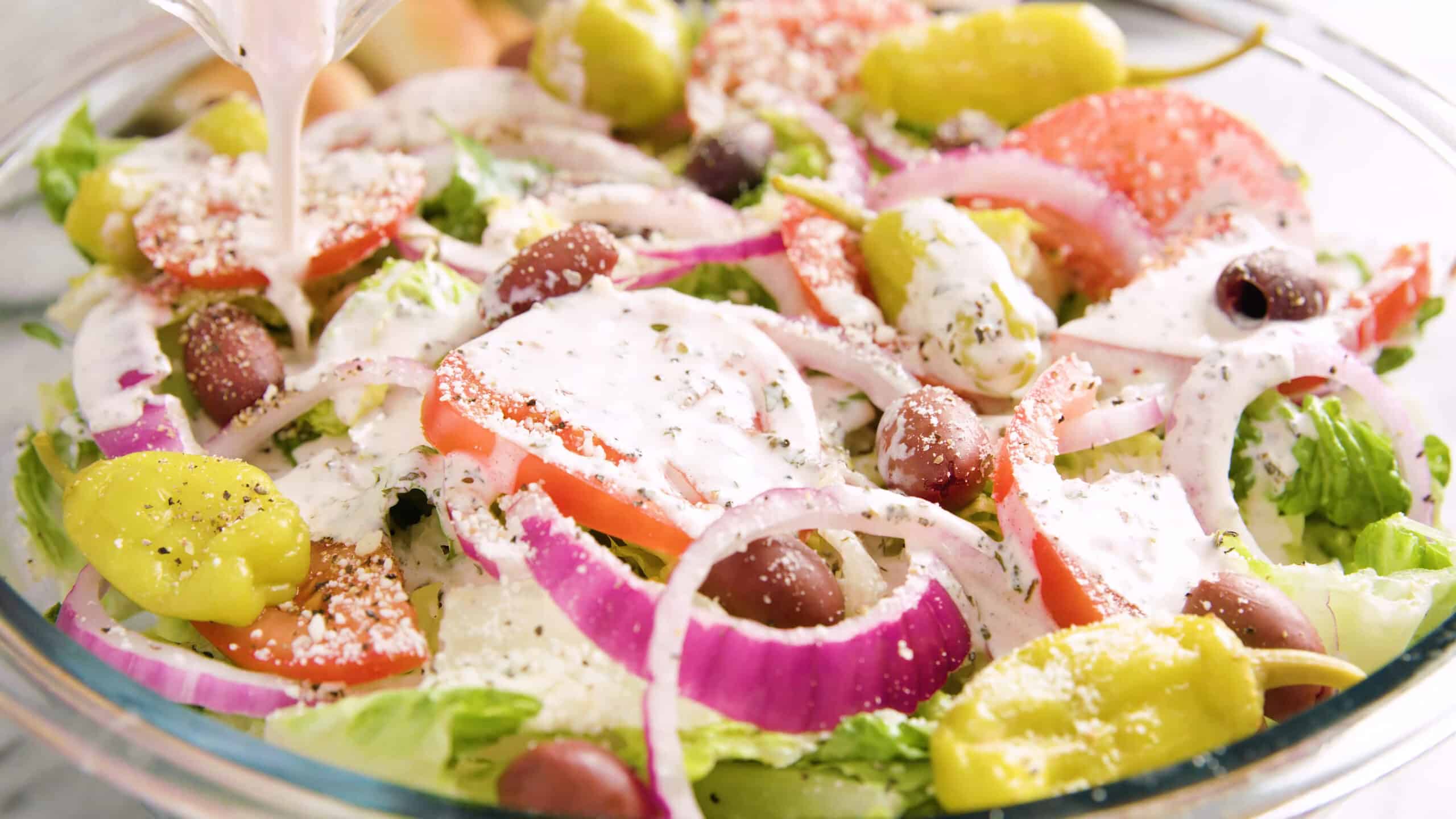 Close-up view of a large clear glass mixing bowl filled to the brim with fresh mixed salad and creamy copycat Olive Garden salad dressing being poured on top.