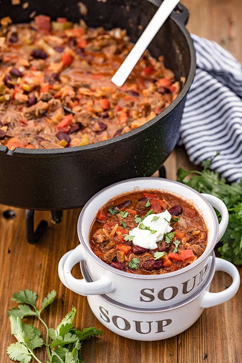  A bowl full of Dutch Oven Chili stack on another bowl with a dutch oven full of chili in the background.