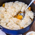 Dutch Oven Peach Cobbler in a blue dutch oven, a scoop has been taken out and put in a bowl