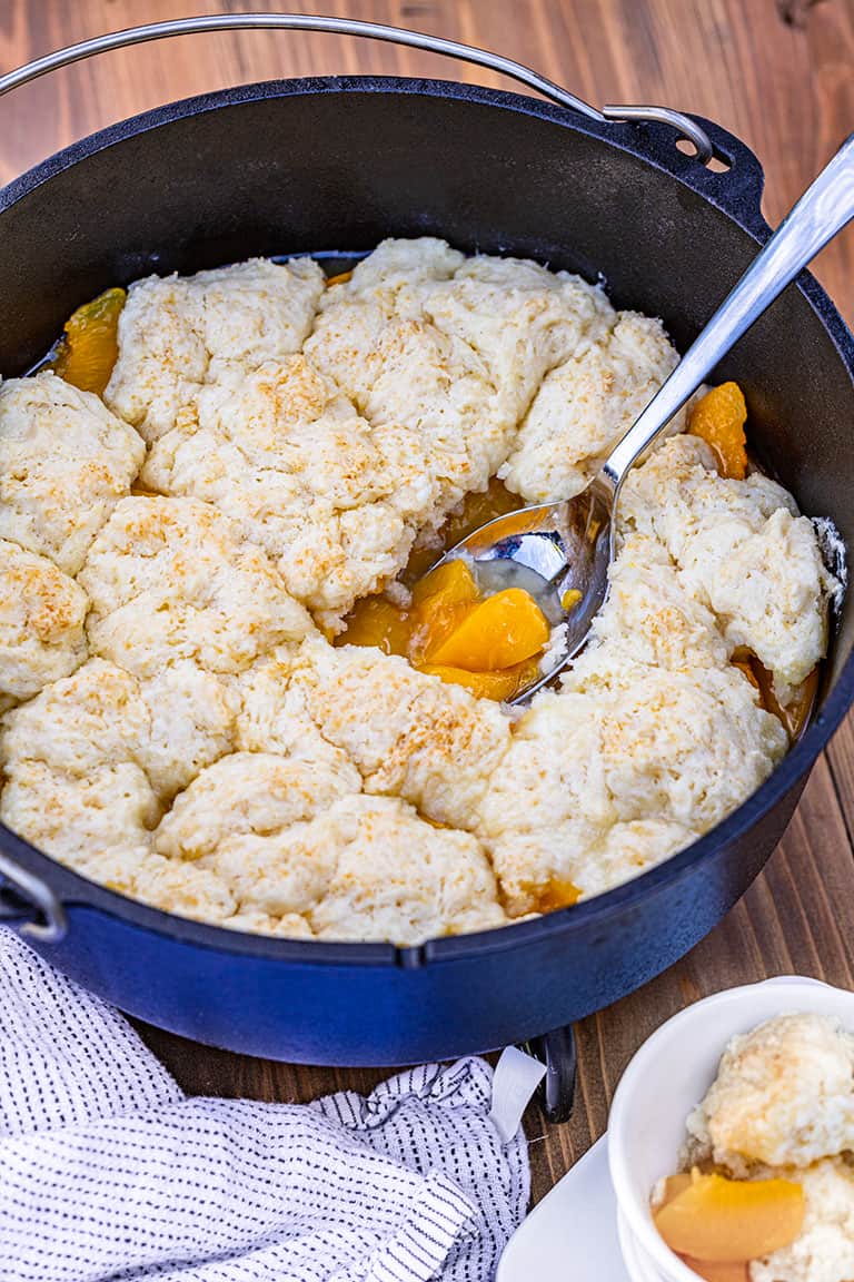 Dutch Oven Peach Cobbler in a blue dutch oven, a scoop has been taken out and put in a bowl