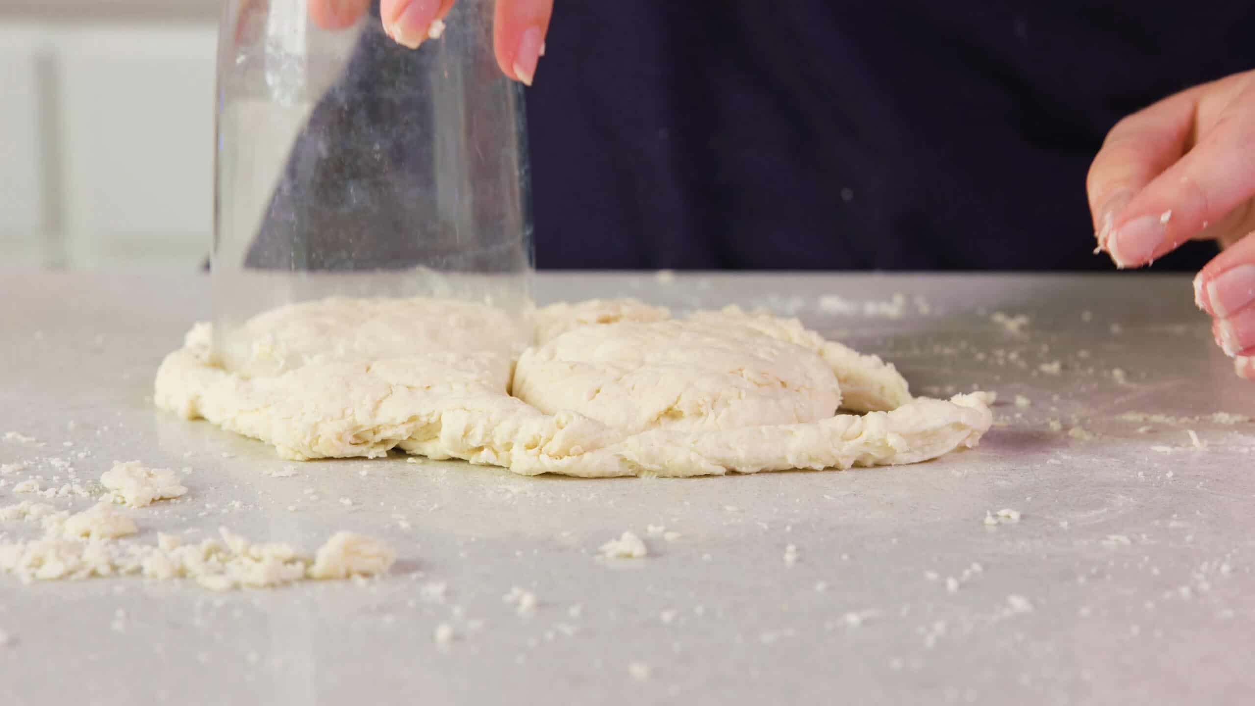 Close-up view of 8 oz glass being used to cut out two individual biscuits from remaining buttermilk biscuit dough on a clean countertop.