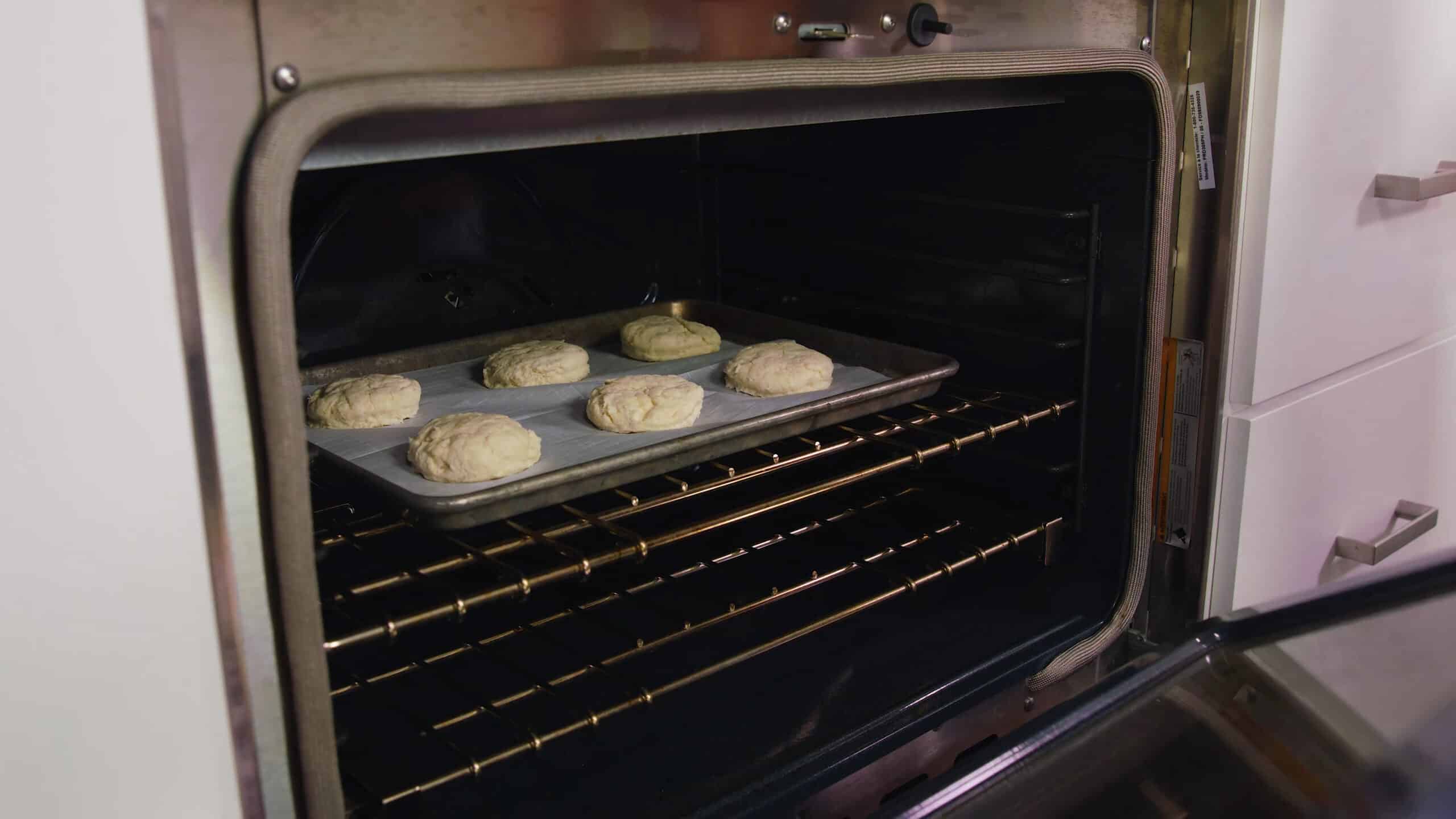 Angled view of interior of an oven with the door open and a baking sheet with parchment paper and six buttermilk biscuits on top.