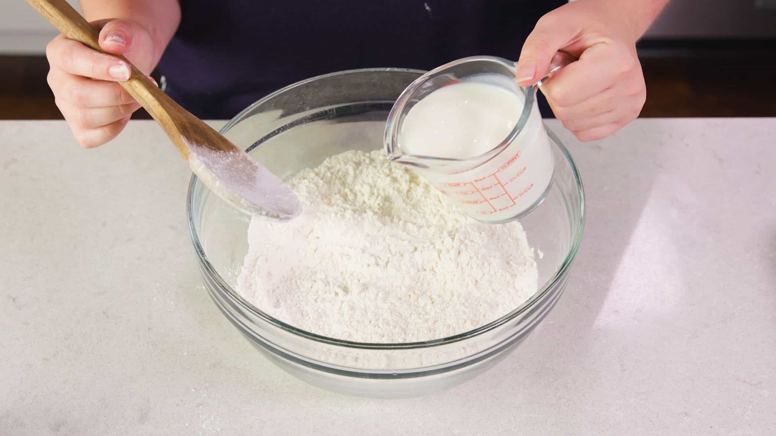 Angled view of mixed ingredients in a mixing bowl on countertop with a wooden spoon in hand and a measuring cup of buttermilk in the other hand ready to pour.