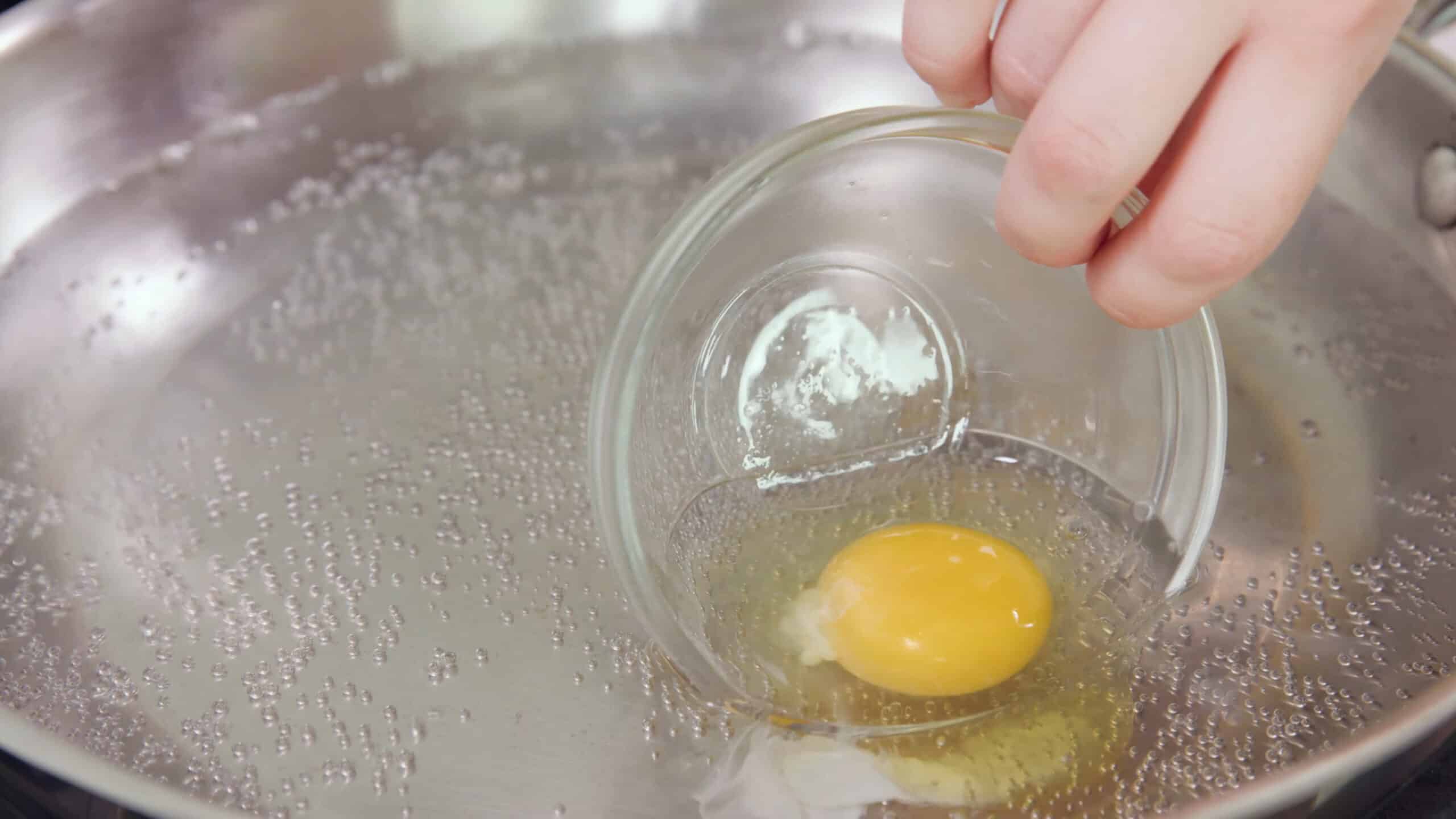 Close-up view of silver saucepan with heated water and a individual egg added to water by tipping small dish.
