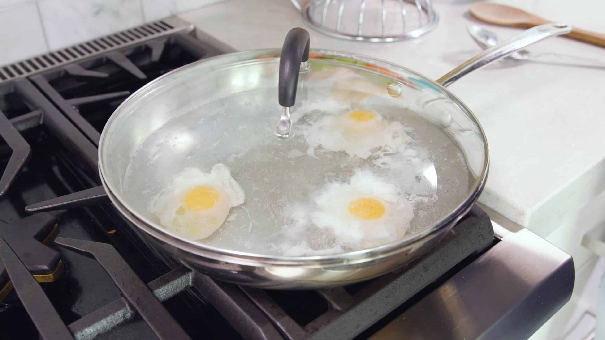 Angled view of silver saucepan with glass lid and three eggs being poached inside on stovetop.