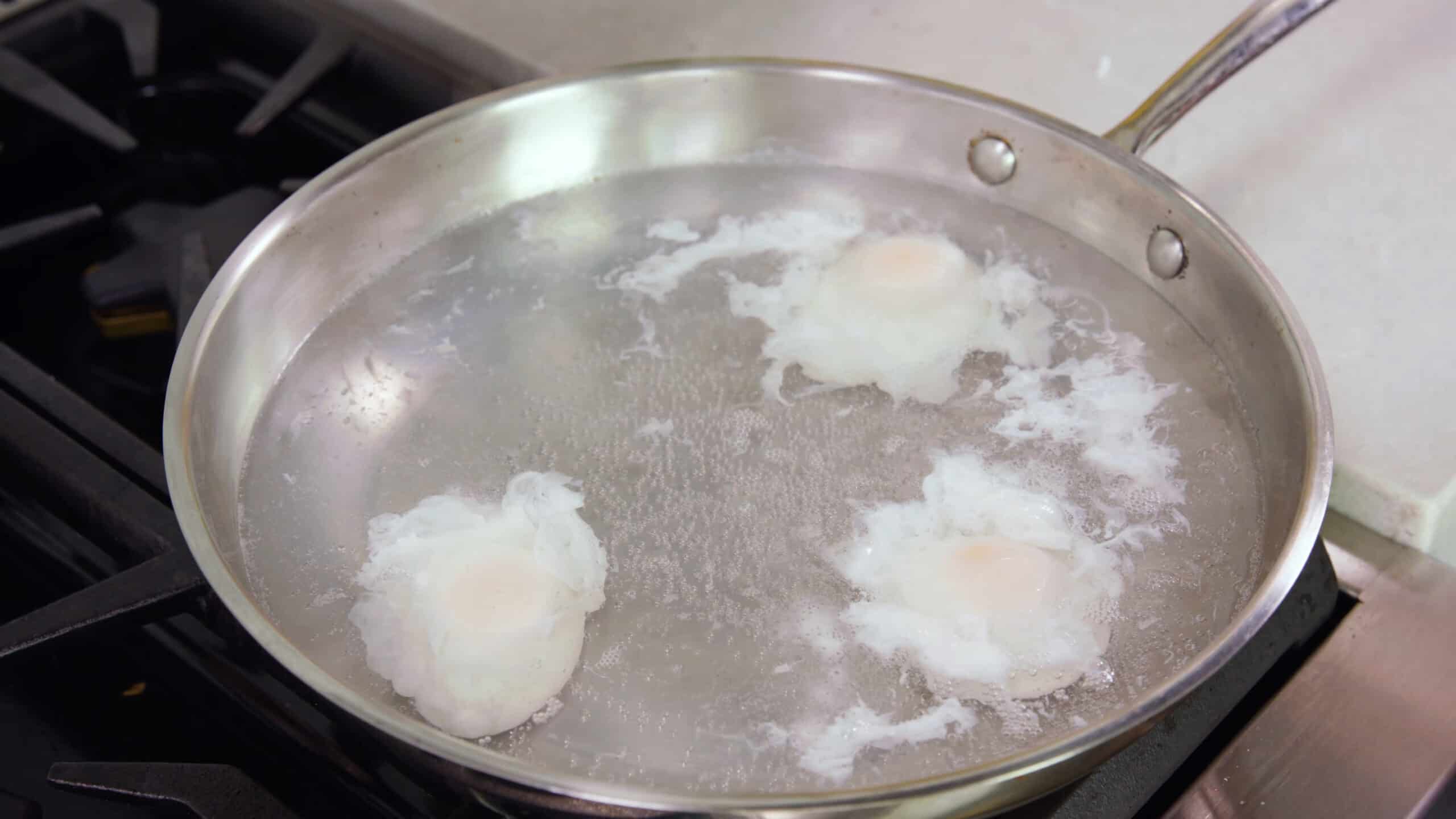Angled view of silver saucepan with three poached eggs inside on stovetop.