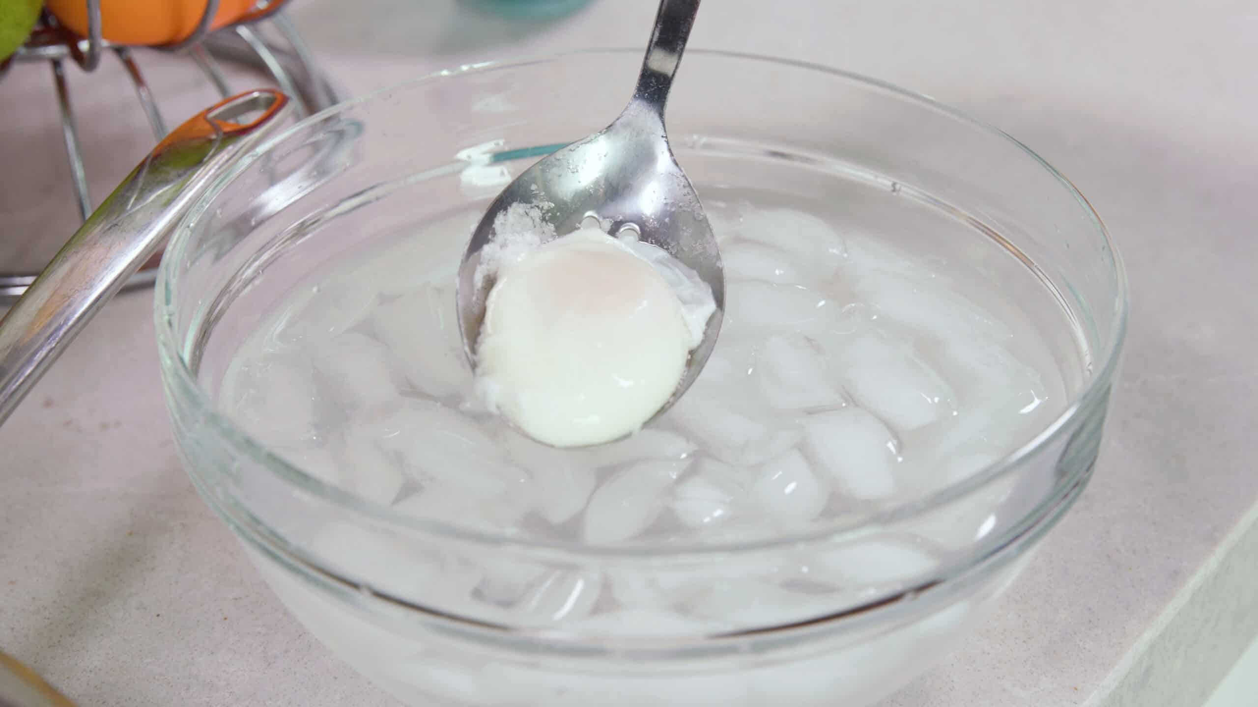Angled view of slotted metal spoon with a single poached egg being placed into an ice bath in a clear mixing bowl on a countertop to cool