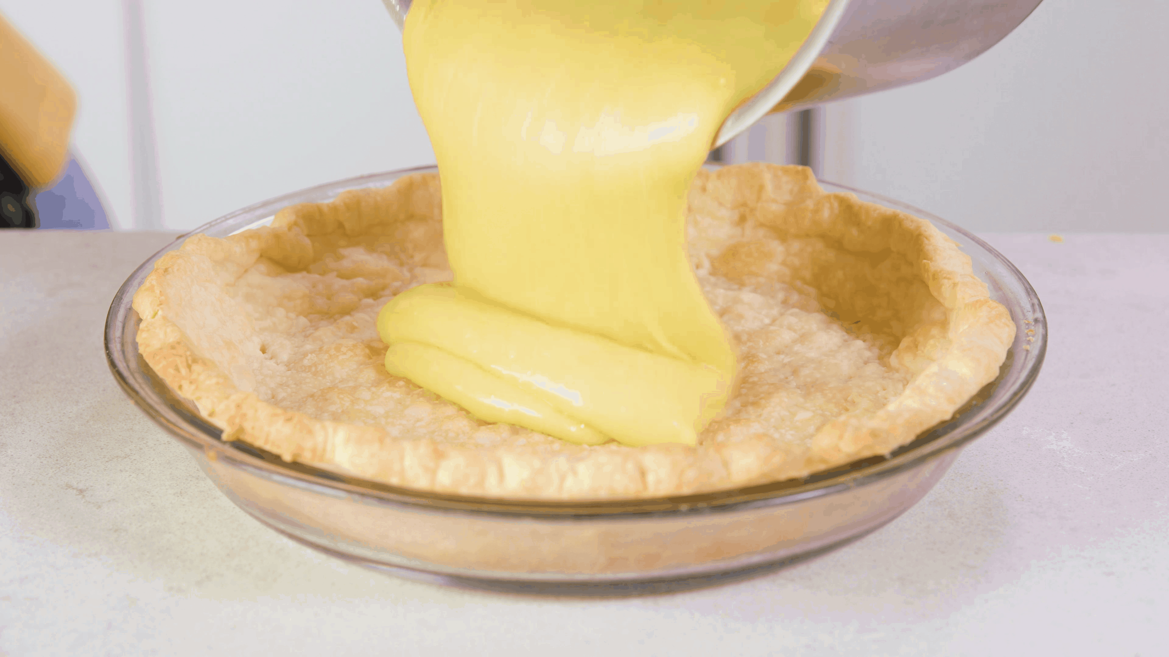 Angled view of prepared pie crust in clear pie plate on countertop and a silver pot pouring in the yellow creamy lemon pie filling.