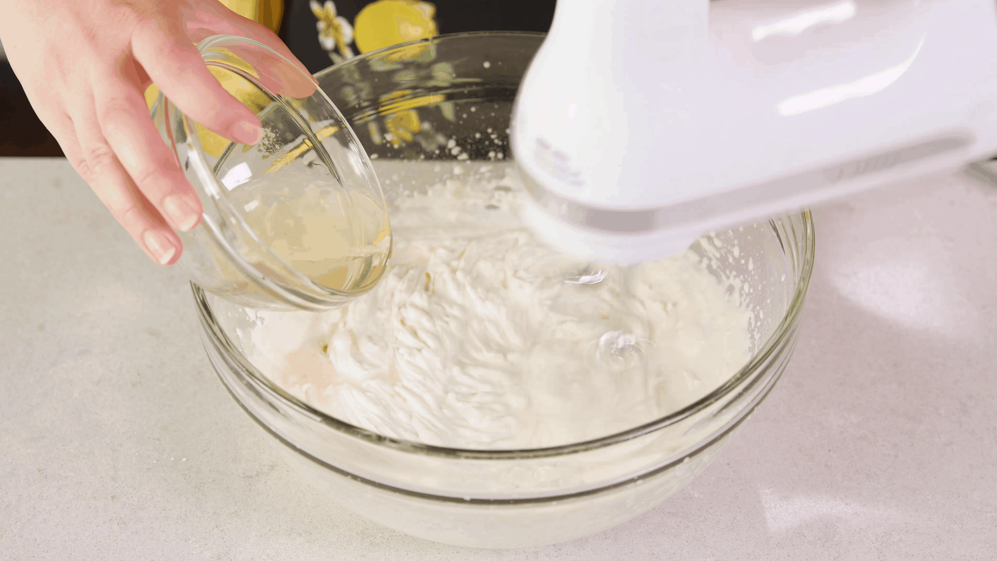 A large mixing bowl with hand mixer whipping cream and a small dish with gelatin being poured from the side to stabilize the whipped cream.