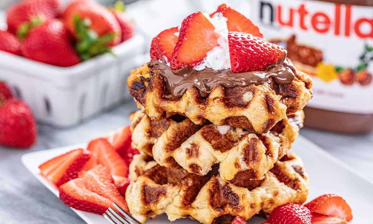 Stack of three Liège Waffles topped with hazelnut chocolate spread, fresh strawberry slices, and whipped cream all on a square white plate.