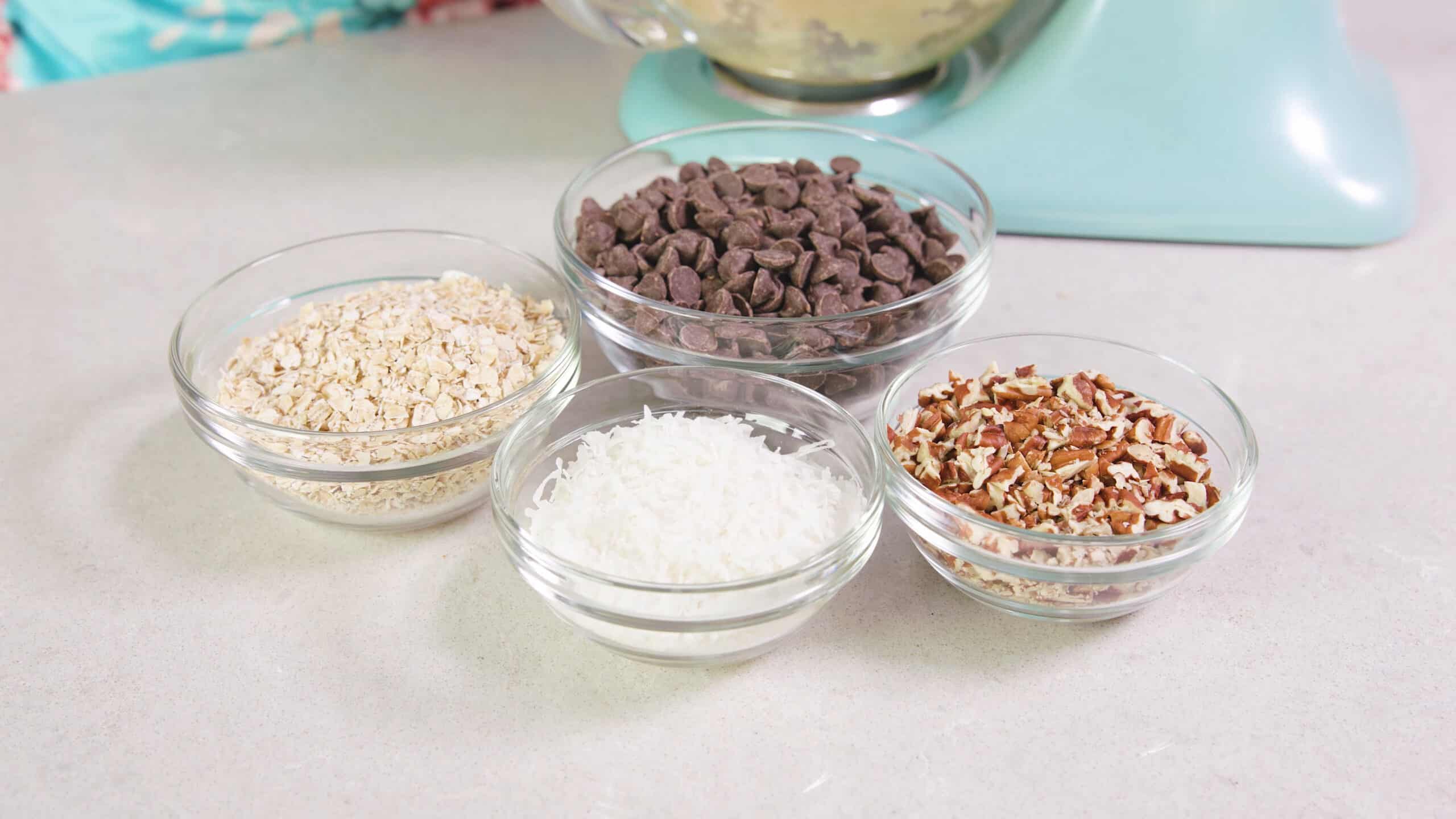 Angled view of small clear glass mixing bowls holding various ingredients to add to the cookie dough like dry oatmeal on the left, sweetened coconut, semisweet chocolate chips and chopped pecans.
