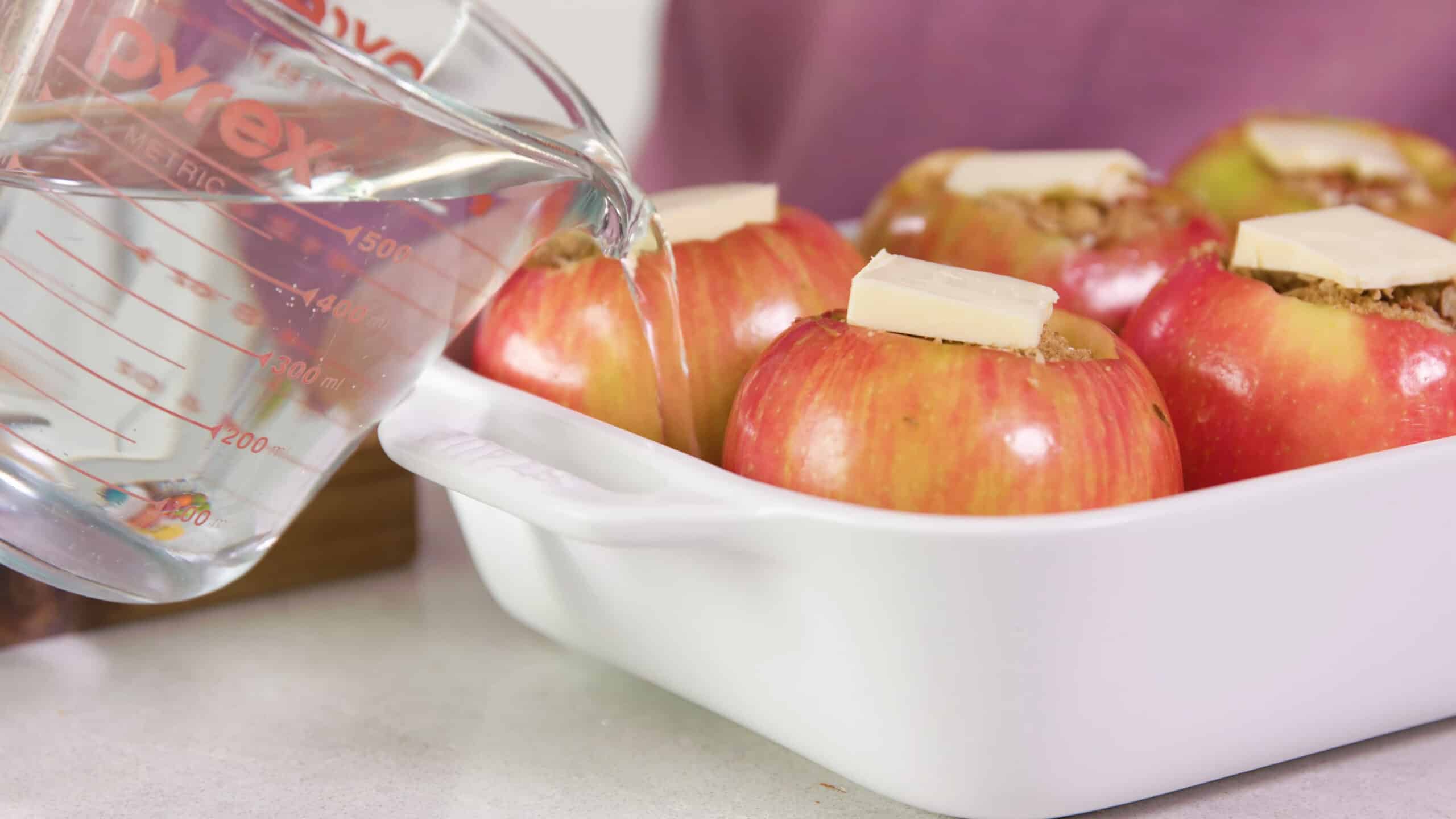 Side view of white serving dish with six apples with filling and a pad of butter on each one and a clear measuring cup full of water being poured in with the apples.