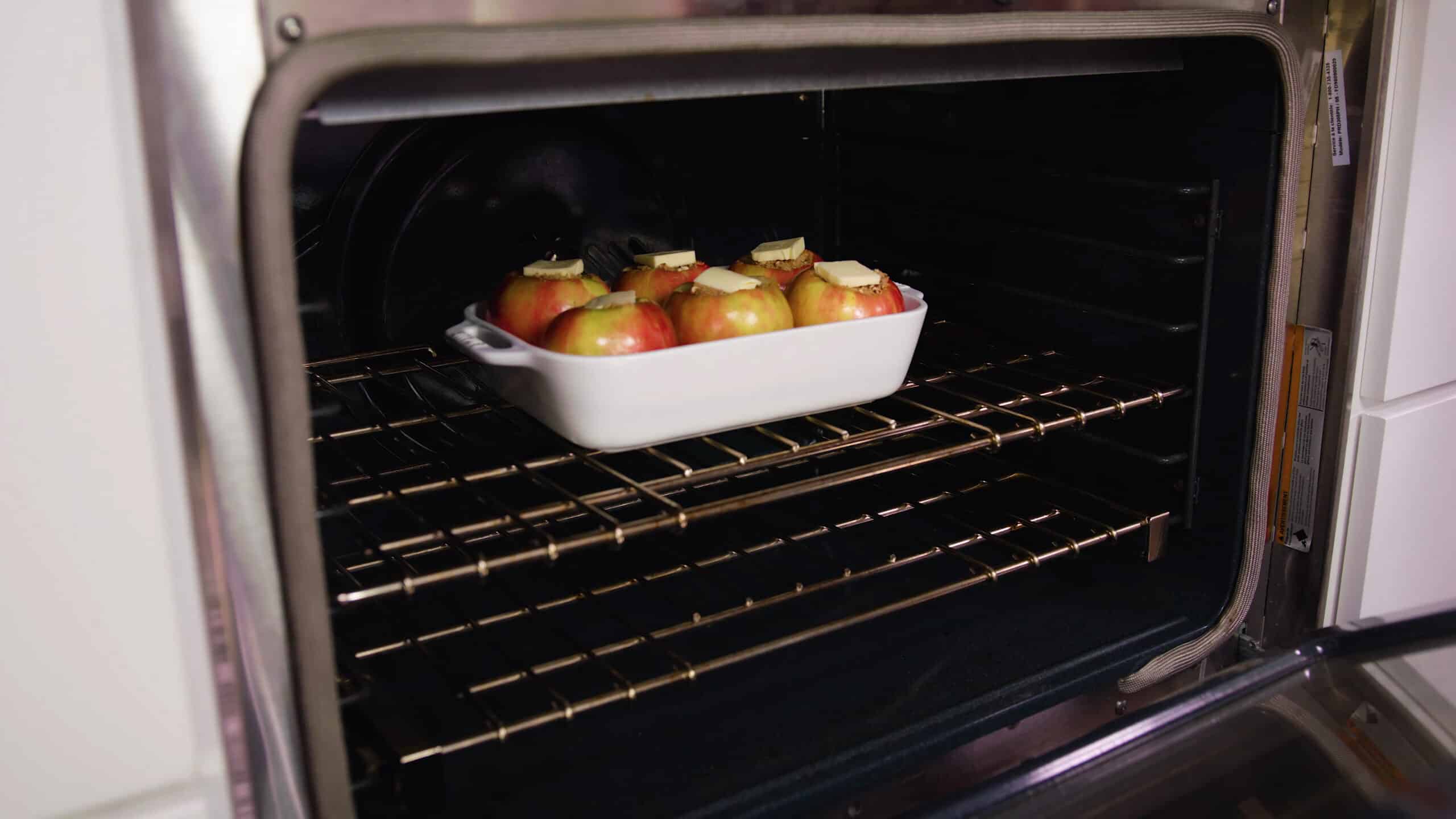 Angled view of open oven with white baking dish with the baked apples and filling with a pad of butter on each.