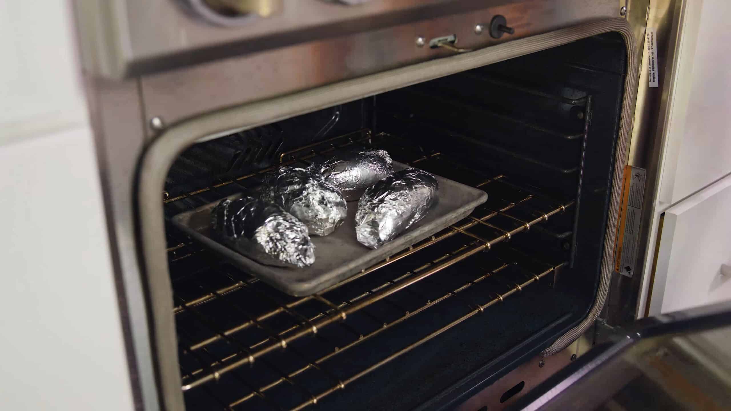 Angled view of four sweet potatoes wrapped in aluminum foil, placed on a baking sheet and put into an open oven.