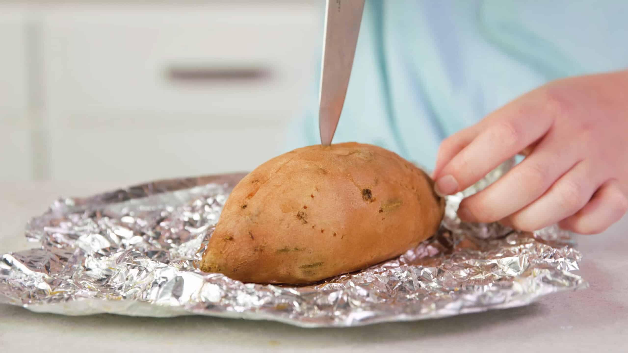 Angled view of one unwrapped baked sweet potato getting sliced down the middle parallel to the ends with a silver kitchen knife.