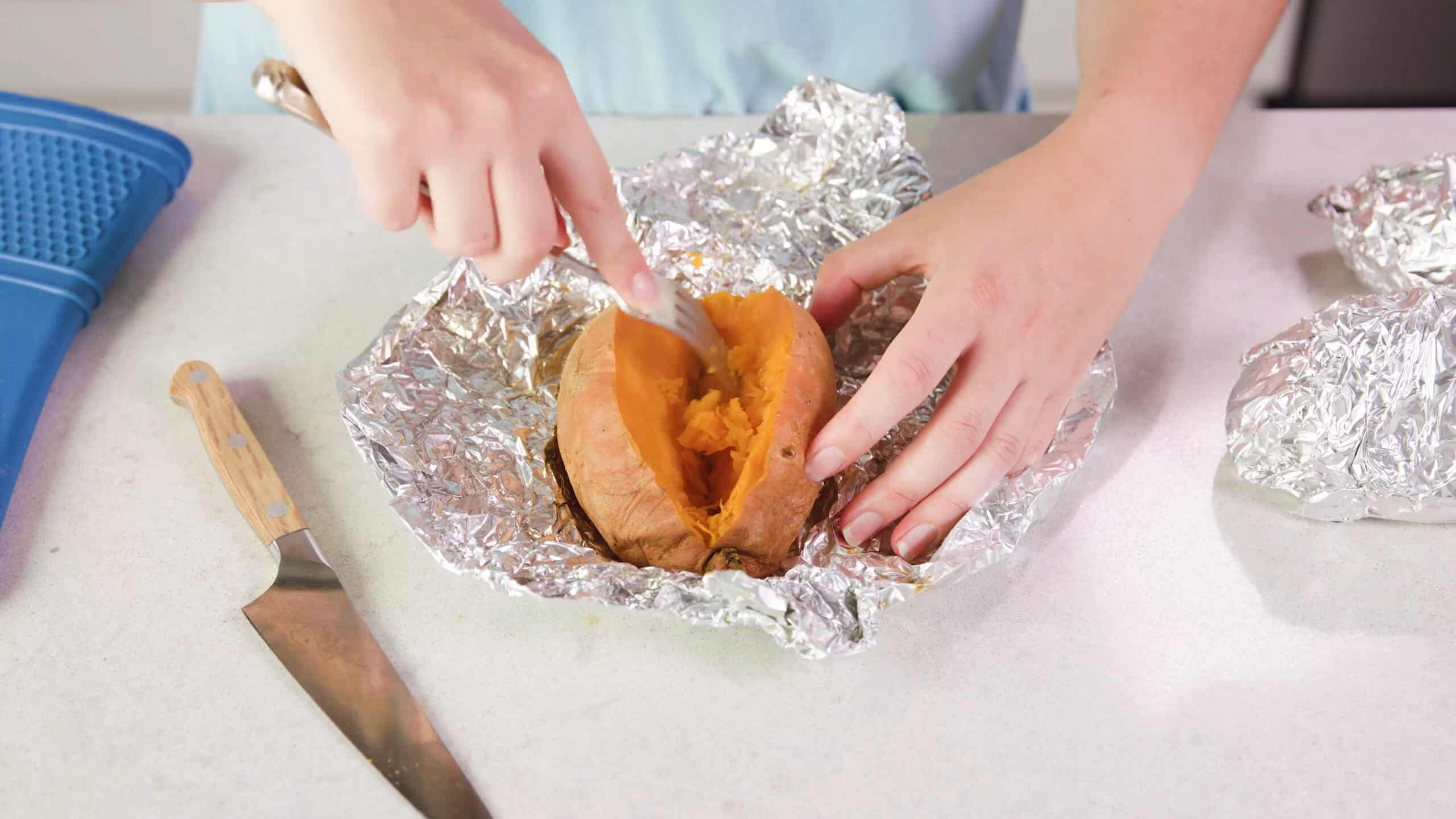 Angled view of the sliced open baked sweet potato on the open aluminum foil and using a silver fork to pull and mash away the interior of the potato.