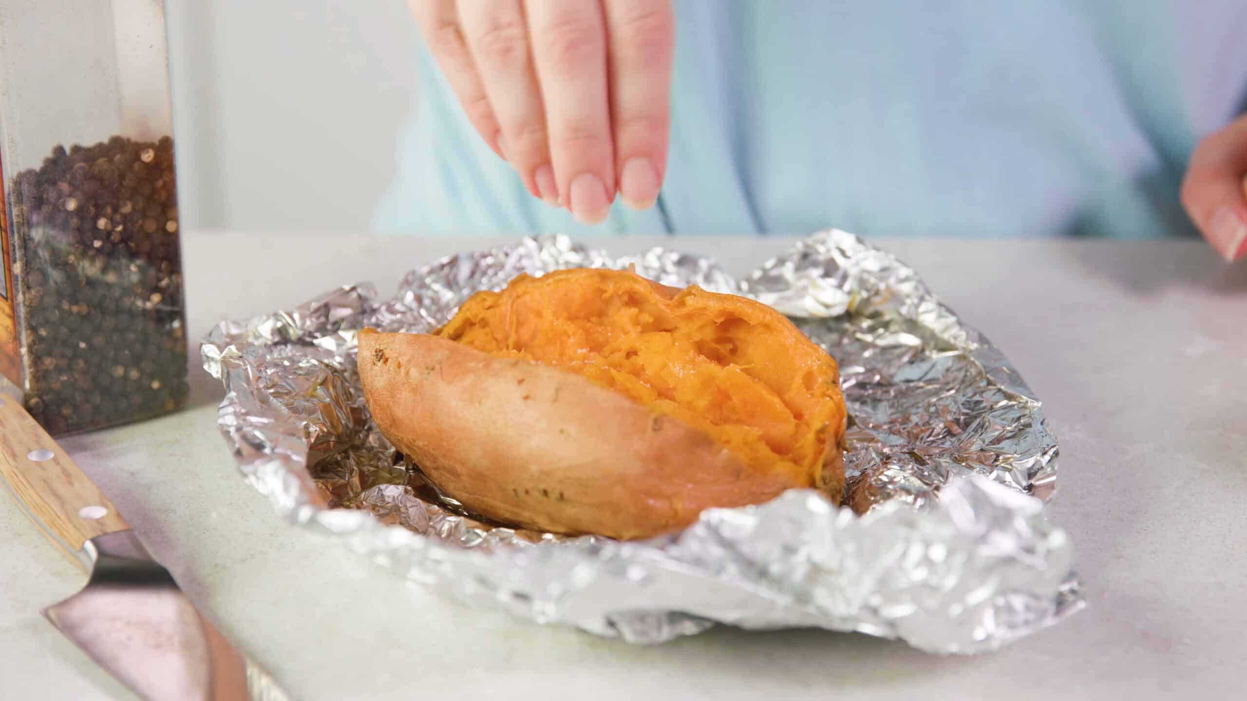 Angled view of open and smashed baked sweet potato on unwrapped aluminum foil with a hand delivering a pinch of salt to the interior of the potato.