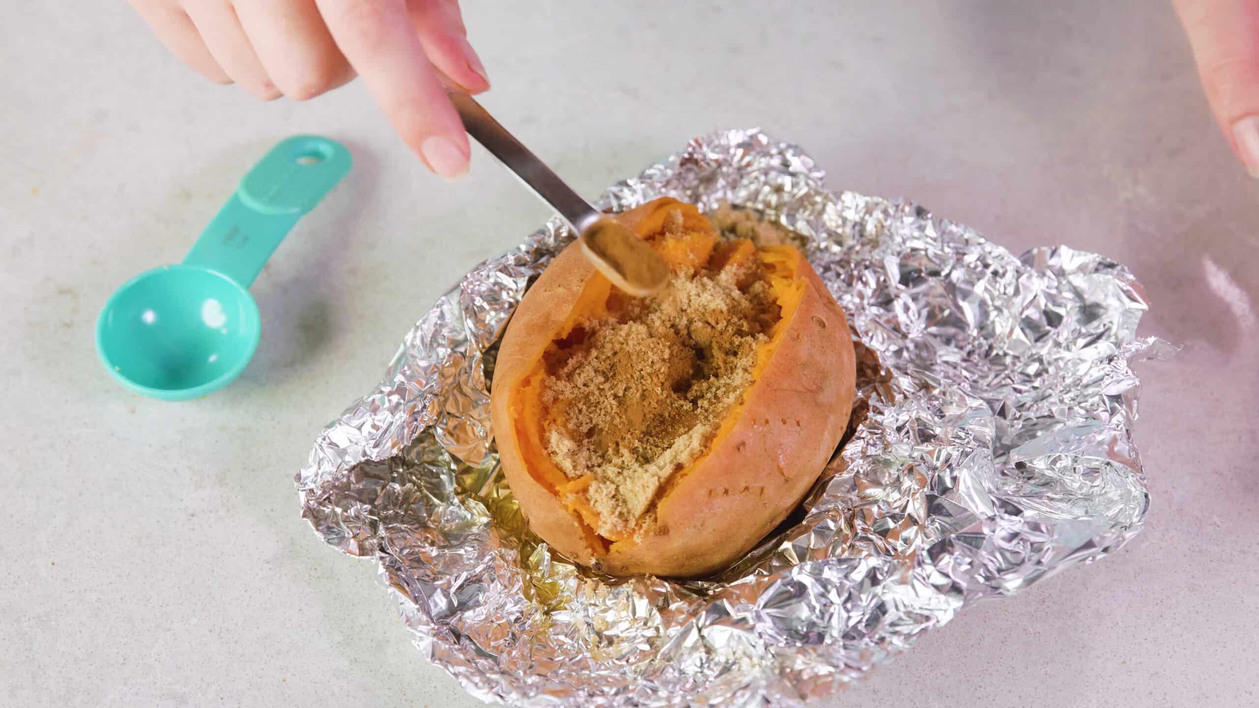 Overhead view an open smashed baked sweet potato with brown sugar being seasoned with cinnamon, all on aluminum foil wrapping on marble countertop.