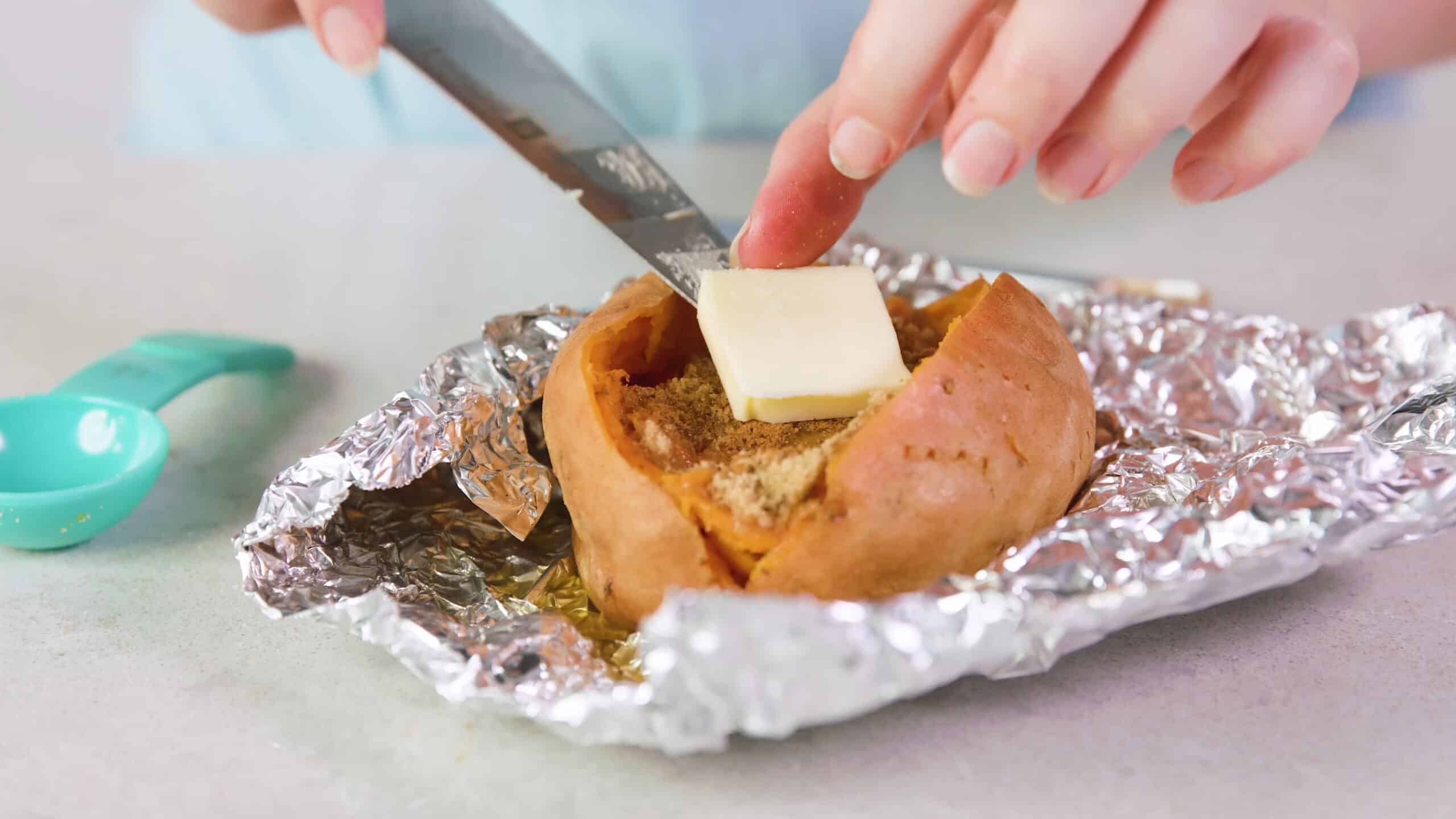 Close-up view of an open smashed baked sweet potato seasoned with brown sugar and cinnamon, and a pad of butter being placed on top with a silver kitchen knife all on a wrapping of aluminum foil on a marble countertop.