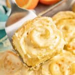 Orange Roll being held up by a metal spatula above a pan full of orange rolls.